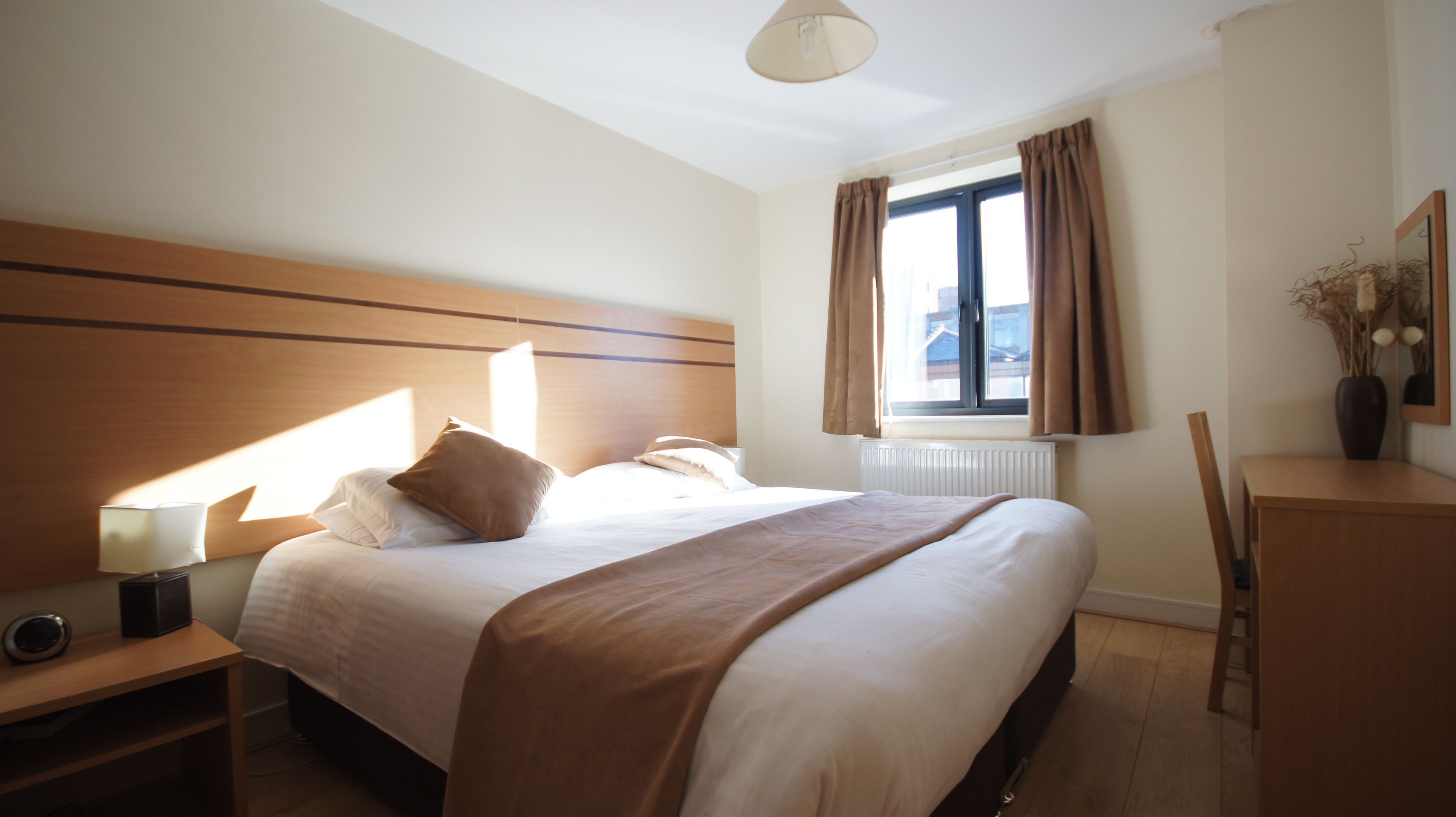 A hotel room with a large bed, a bedside table with a lamp, a chair, a desk, and a window with curtains.