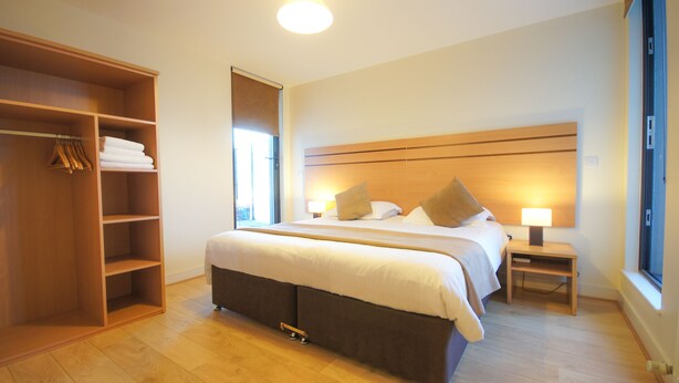 A hotel room with a large bed, a wardrobe, a bedside table with a lamp, and a window. - Crompton Court (London)