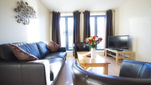A living room with a sofa, a coffee table with a vase of flowers, a television on a stand, and a window with curtains. - Crompton Court (London)
