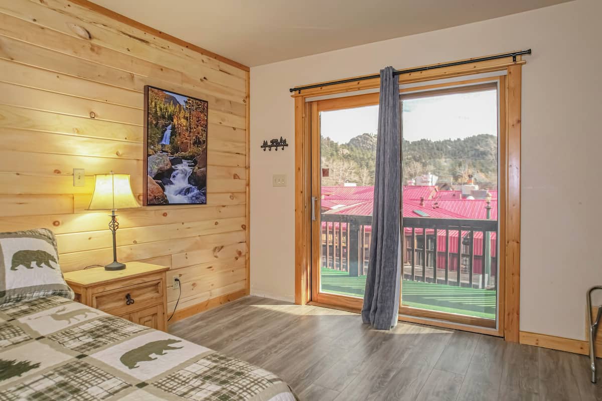 A wooden-paneled bedroom with a bed, a nightstand, a lamp, a painting of a waterfall, and a sliding glass door leading to an outdoor area with a red-roofed building.