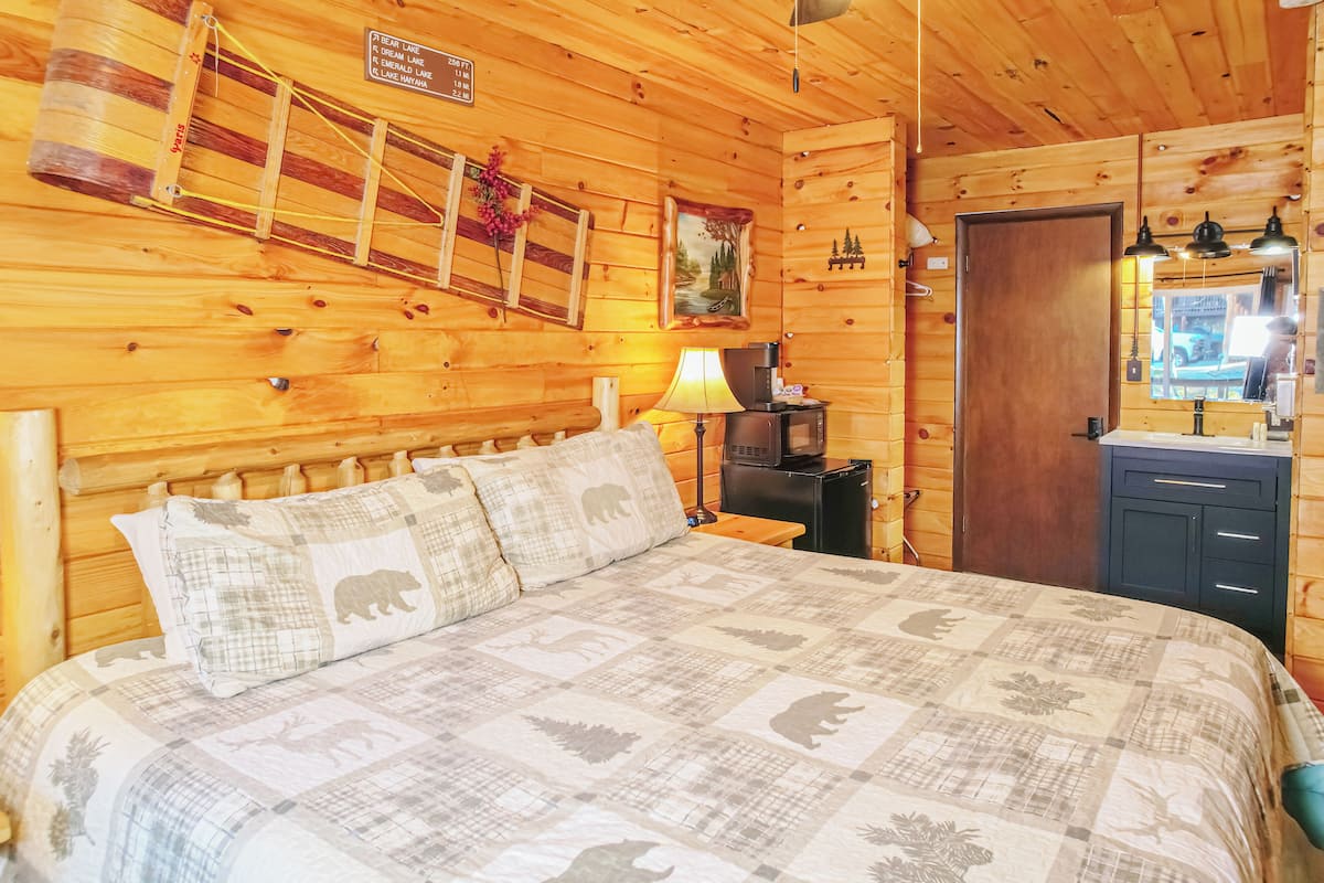 A wooden cabin bedroom with a bed, a nightstand, a lamp, a dresser, and a mirror.