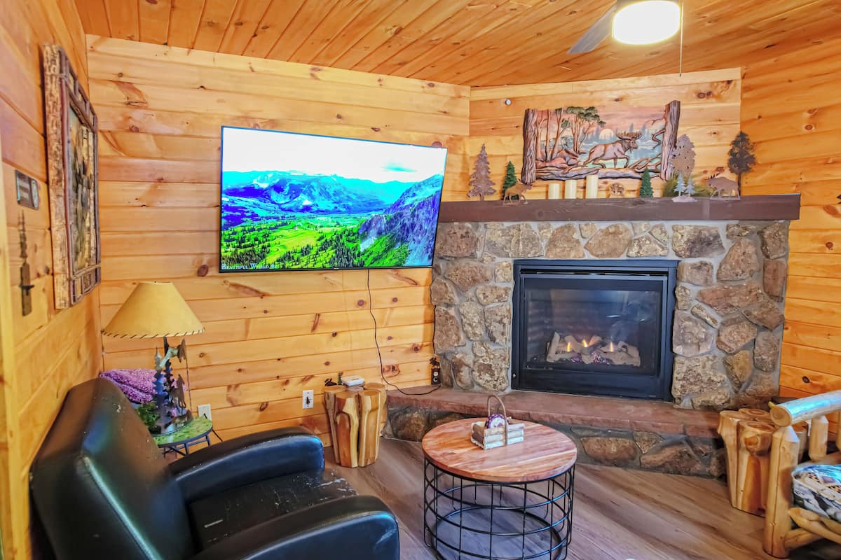 A wooden cabin interior with a stone fireplace, a television mounted on the wall, and a cozy seating area.