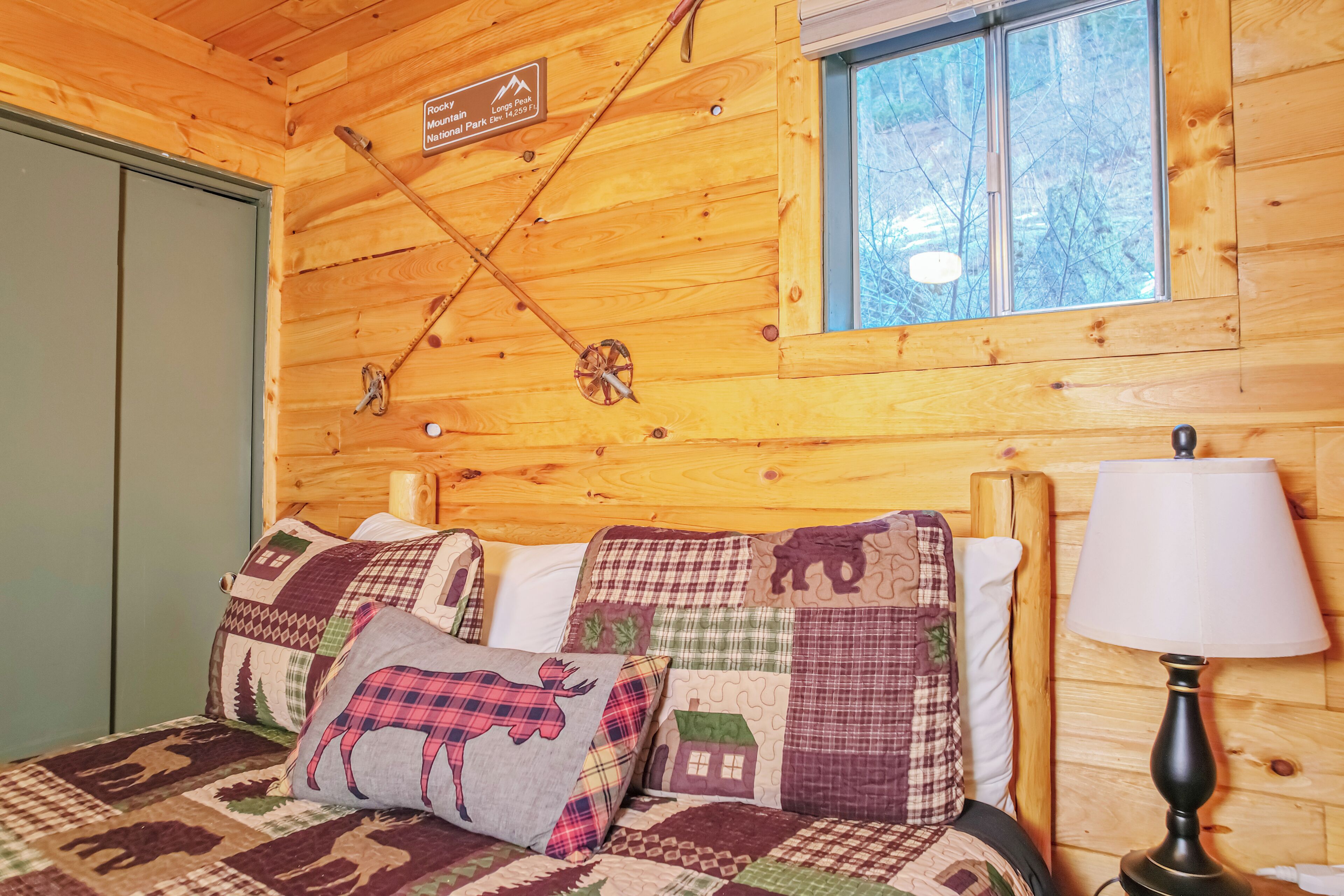 A wooden cabin room with a bed, pillows, a lamp, and a window with a view of trees.