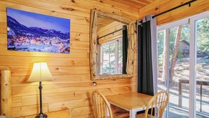 A wooden cabin interior with a dining table, chairs, a lamp, a framed picture, and a window with curtains. - Nicky's Resort (Estes Park)