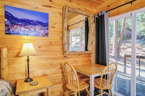 A wooden cabin interior with a dining table, chairs, a lamp, a framed picture, and a window with curtains.