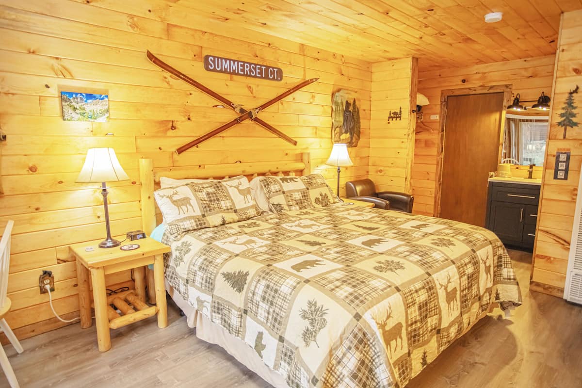 A wooden cabin bedroom with a bed, two bedside lamps, a nightstand, a wooden door, and a wall sign reading 'SUMMERSET CT.'