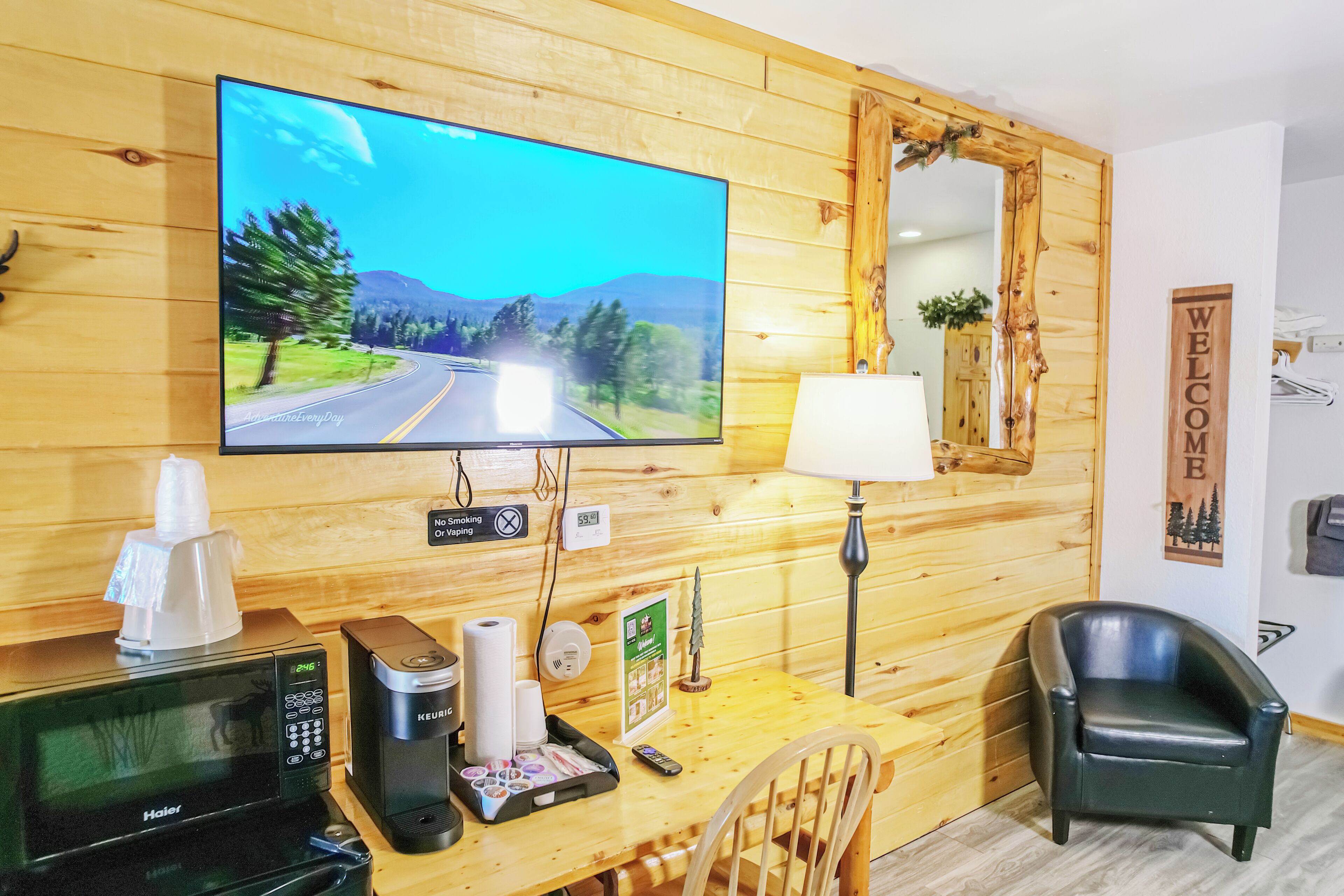 Un interior de madera con una televisión mostrando un paisaje vial, una cafetera, un microondas, una lámpara y un cartel que dice 'WELCOME'.