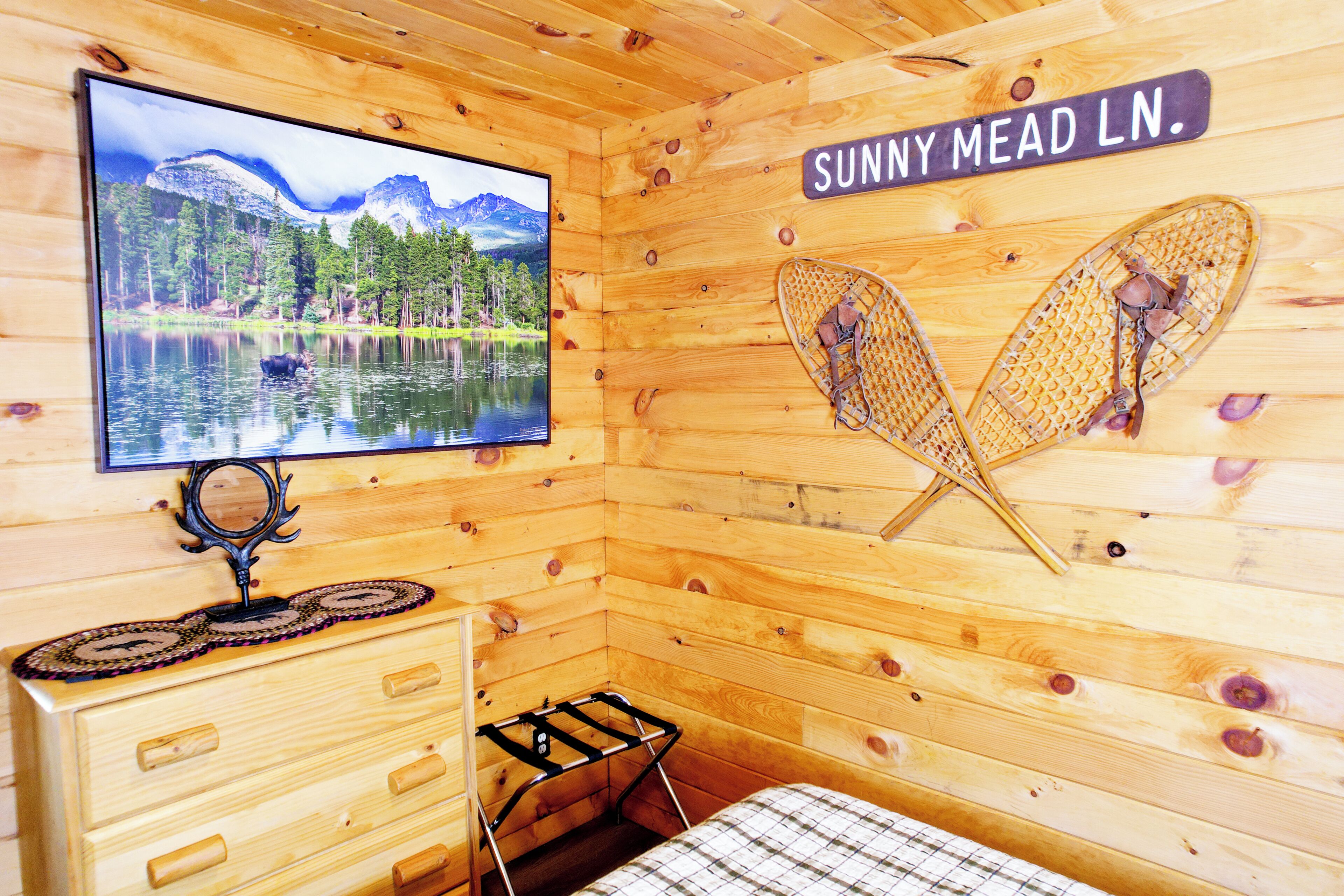 Ein Holzblockhaus-Innenraum mit einem Fernseher, der einen Bergsee zeigt, einem Bett, einem Schminktisch und einem Schild mit der Aufschrift 'Sunny Mead LN'.