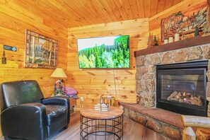 A wooden cabin interior with a fireplace, a television mounted on the wall, a black leather chair, and a small round table. - Nicky's Resort (Estes Park)