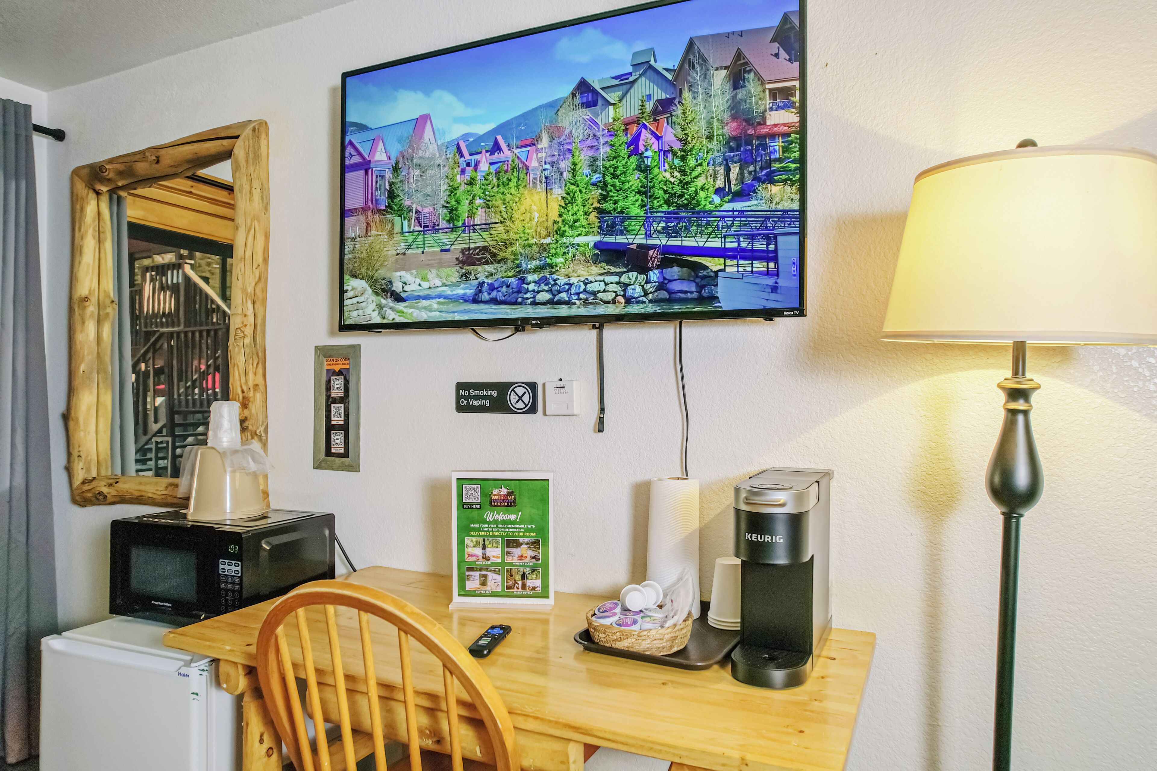 Habitación de hotel con televisión montada en la pared, cafetera, microondas, refrigerador, una mesa de madera y una silla.