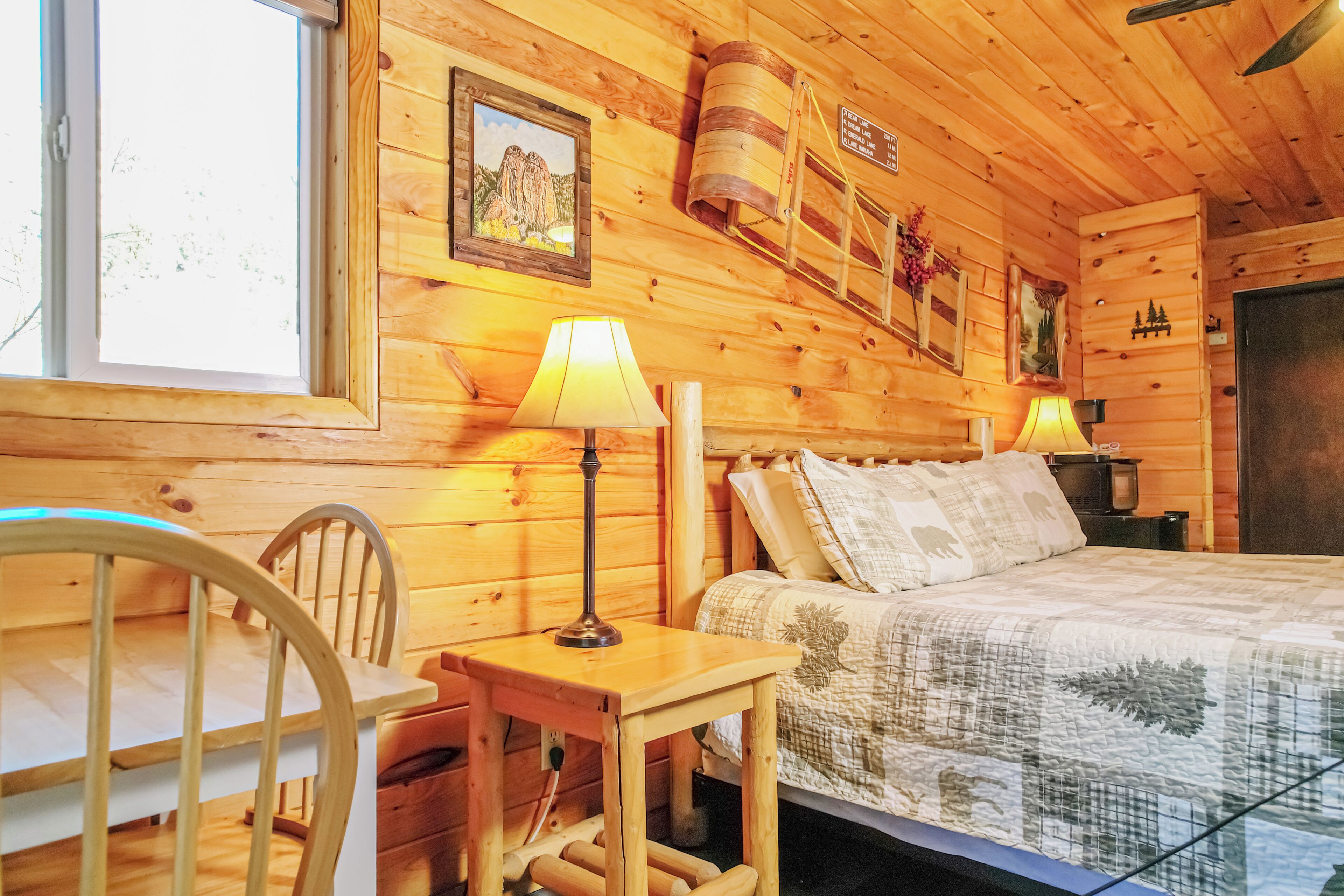 A wooden cabin room with a bed, two lamps, a chair, and a window.
