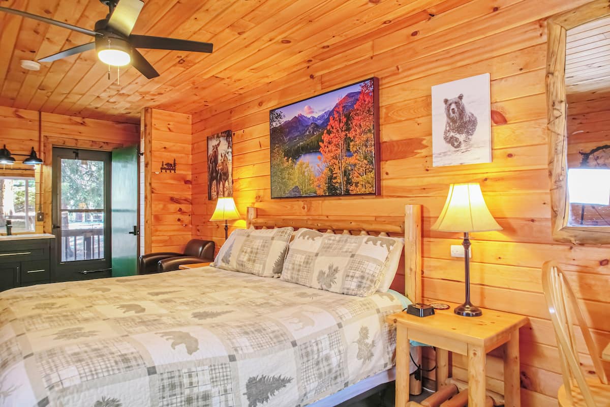 A wooden cabin room with a bed, two wall-mounted lamps, a ceiling fan, and two framed pictures.