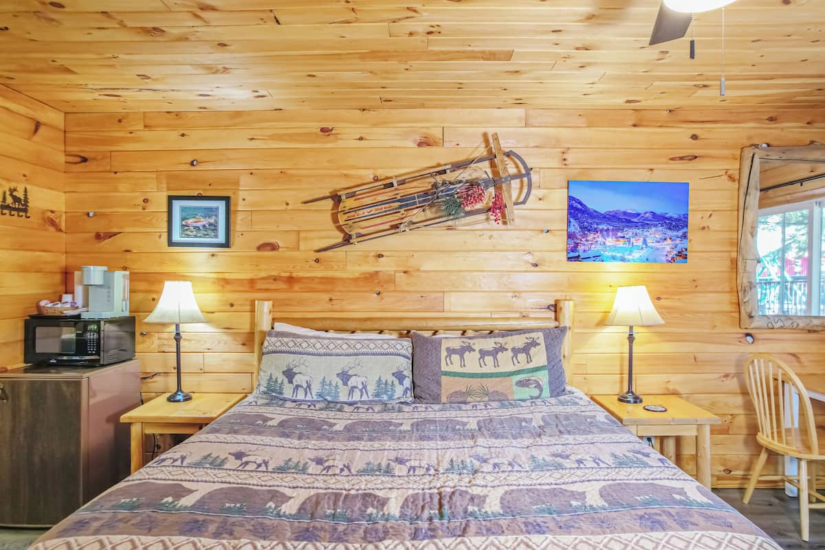 A wooden cabin room with a bed, two nightstands, a desk, a chair, a microwave, and a framed picture on the wall.