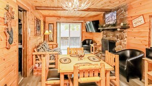 A wooden cabin interior with a dining table, chairs, a stone fireplace, and a television mounted on the wall. - Nicky's Resort (Estes Park)