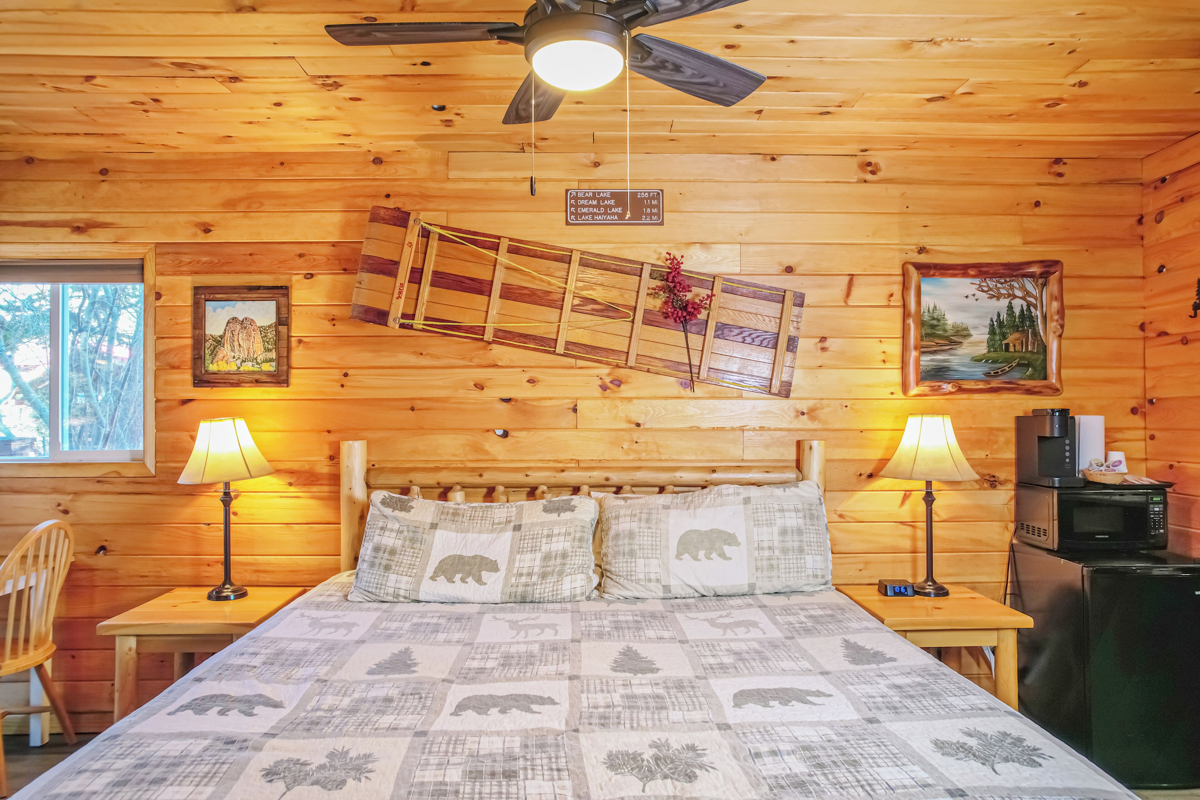 A wooden cabin room with a bed, two nightstands, a ceiling fan, a window, and a wall-mounted sign indicating distances to nearby lakes.