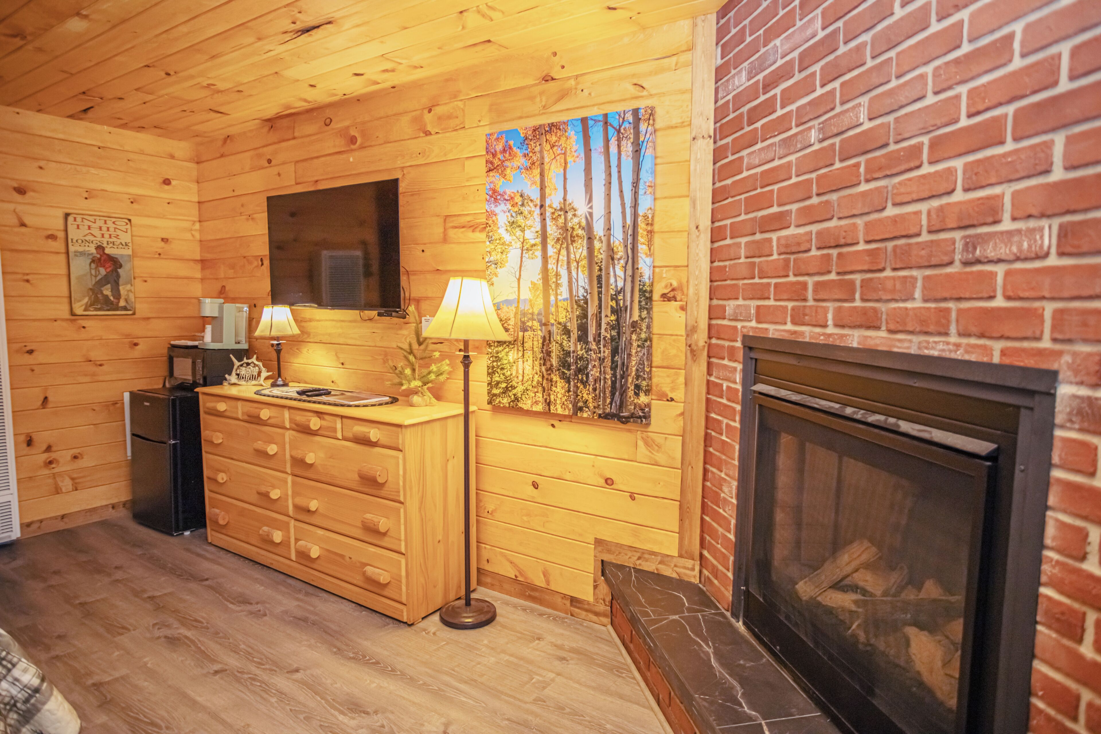 Interior de una cabaña de madera con chimenea, un aparador de madera, un televisor y un cuadro con árboles.