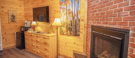 Interior de una cabaña de madera con chimenea, un aparador de madera, un televisor y un cuadro con árboles.