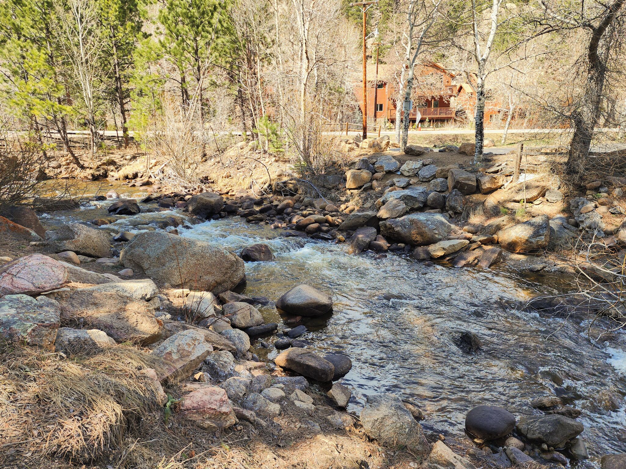 A natural stream with rocks and trees, a wooden bridge, and a cabin in the background.