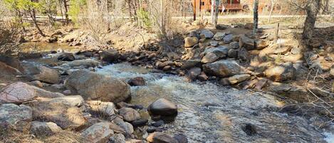 A natural stream with rocks and trees, a wooden bridge, and a cabin in the background.