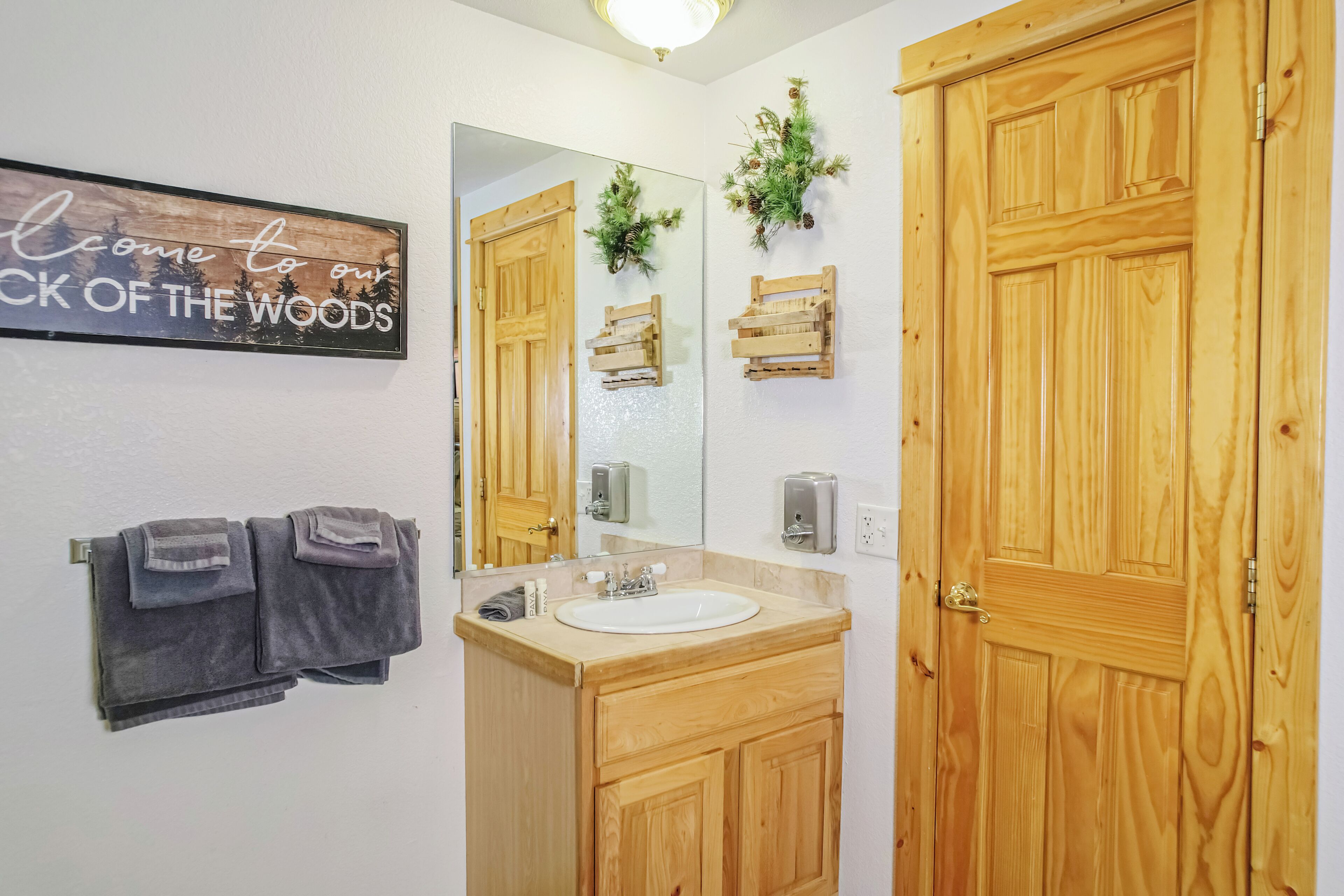Un baño con armarios de madera, un lavabo, un espejo y un letrero que dice 'Welcome to our Back of the Woods'.