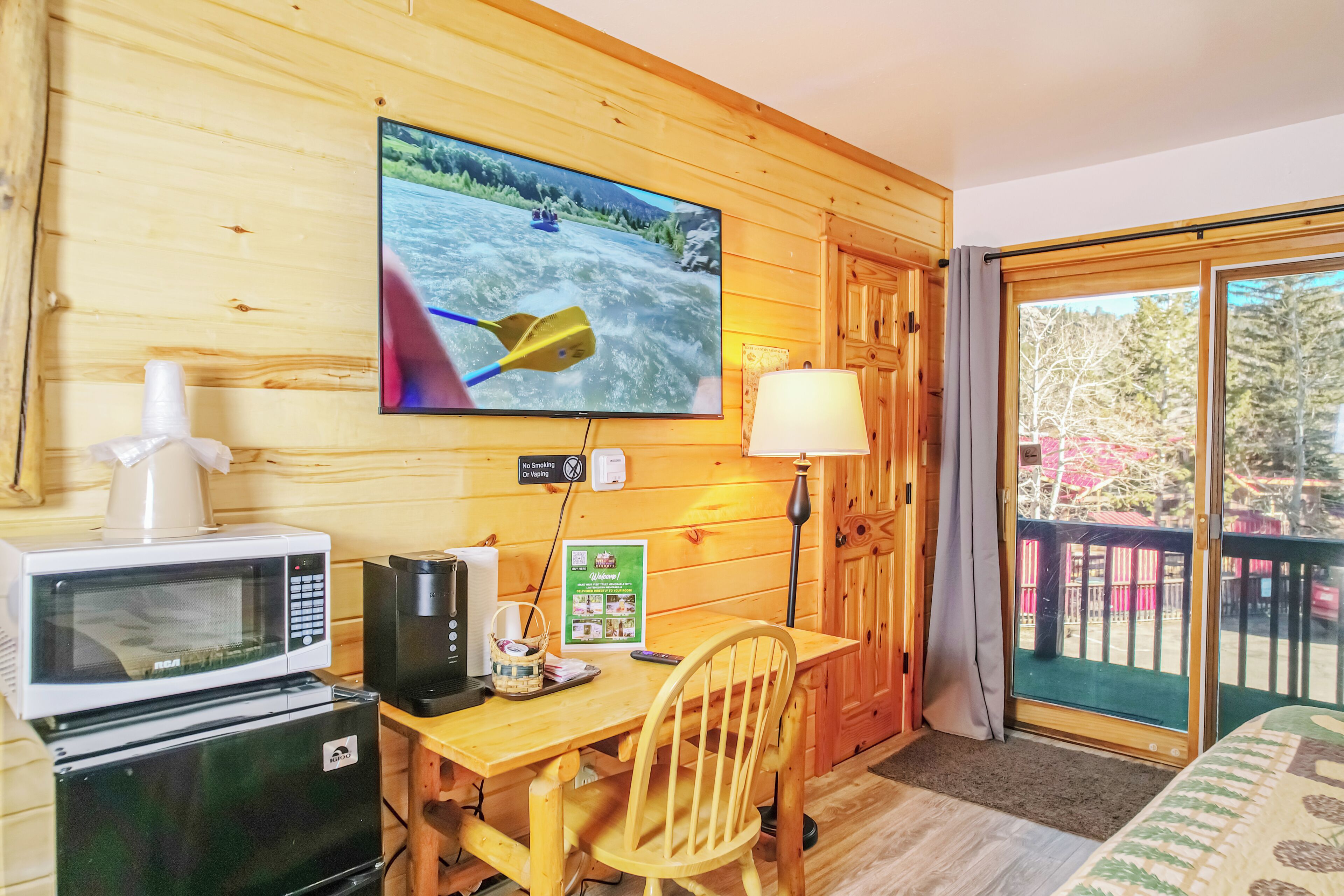 A wooden cabin room with a television, microwave, refrigerator, table, chair, and a view of a forest through a sliding door.