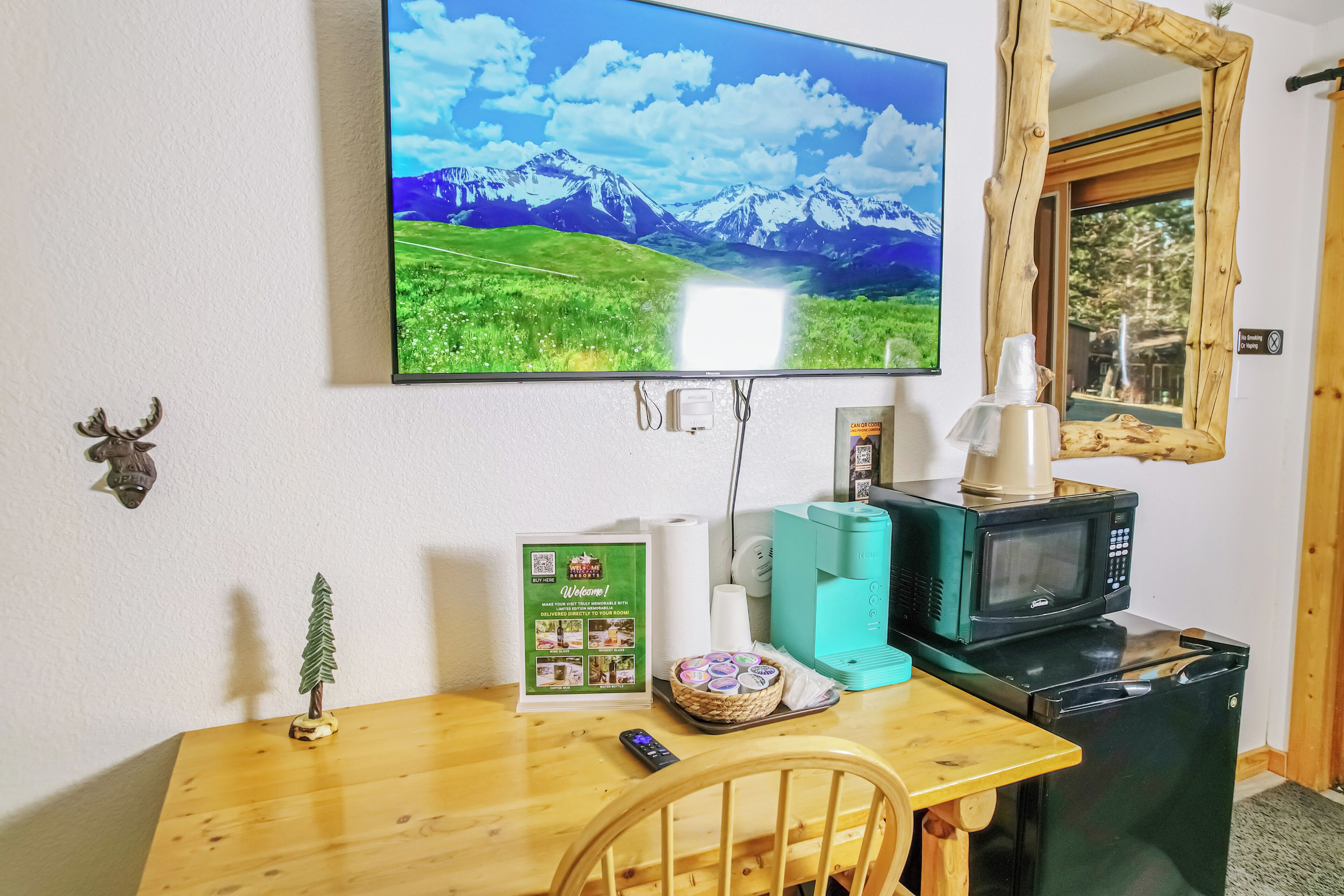 A room with a wooden table, a television displaying a mountain landscape, a small decorative tree, a welcome sign, a microwave, and a refrigerator.