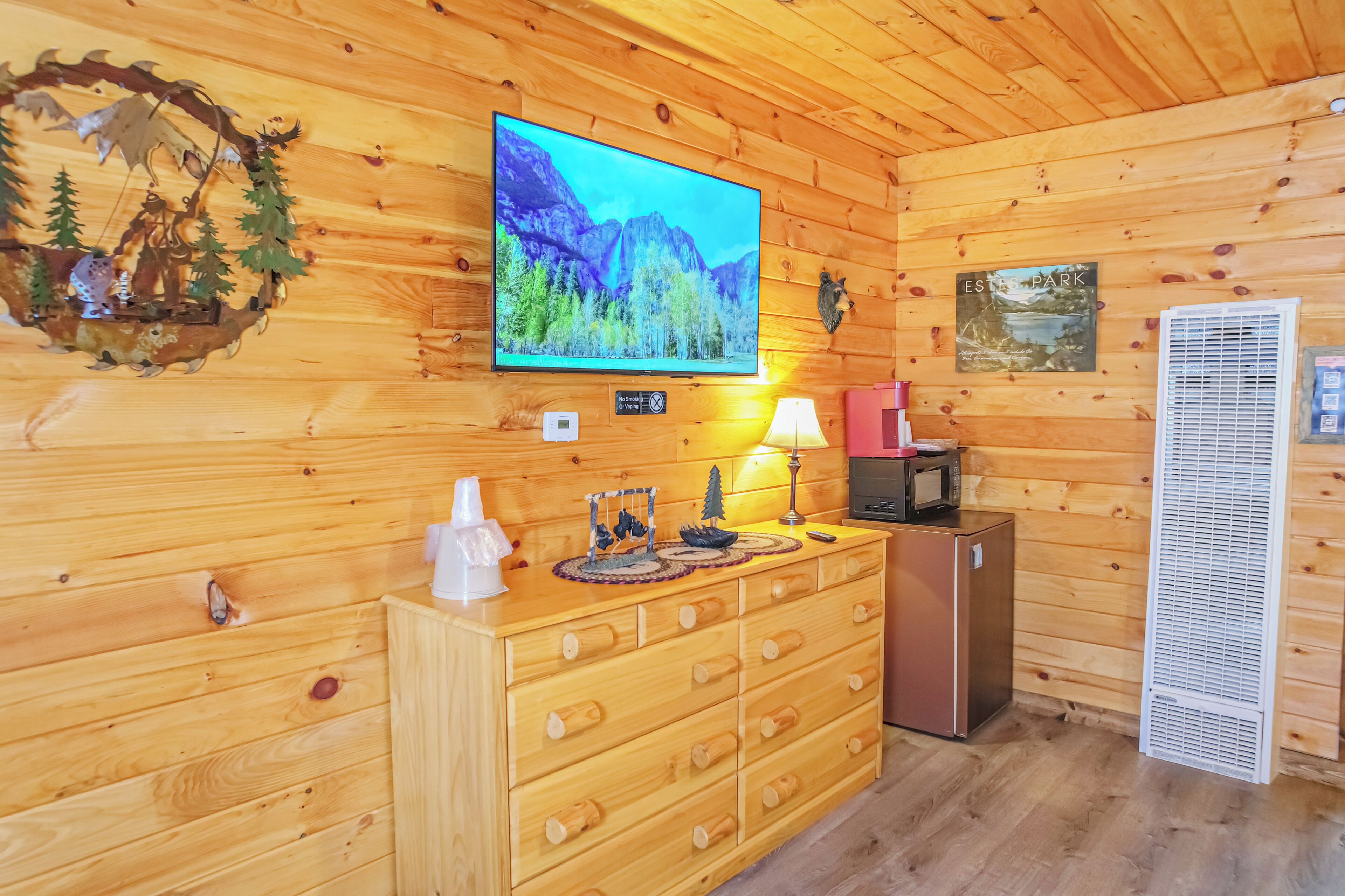Interior de una cabaña de madera con una televisión montada, un aparador de madera, un refrigerador, una lámpara y un calentador de pared.