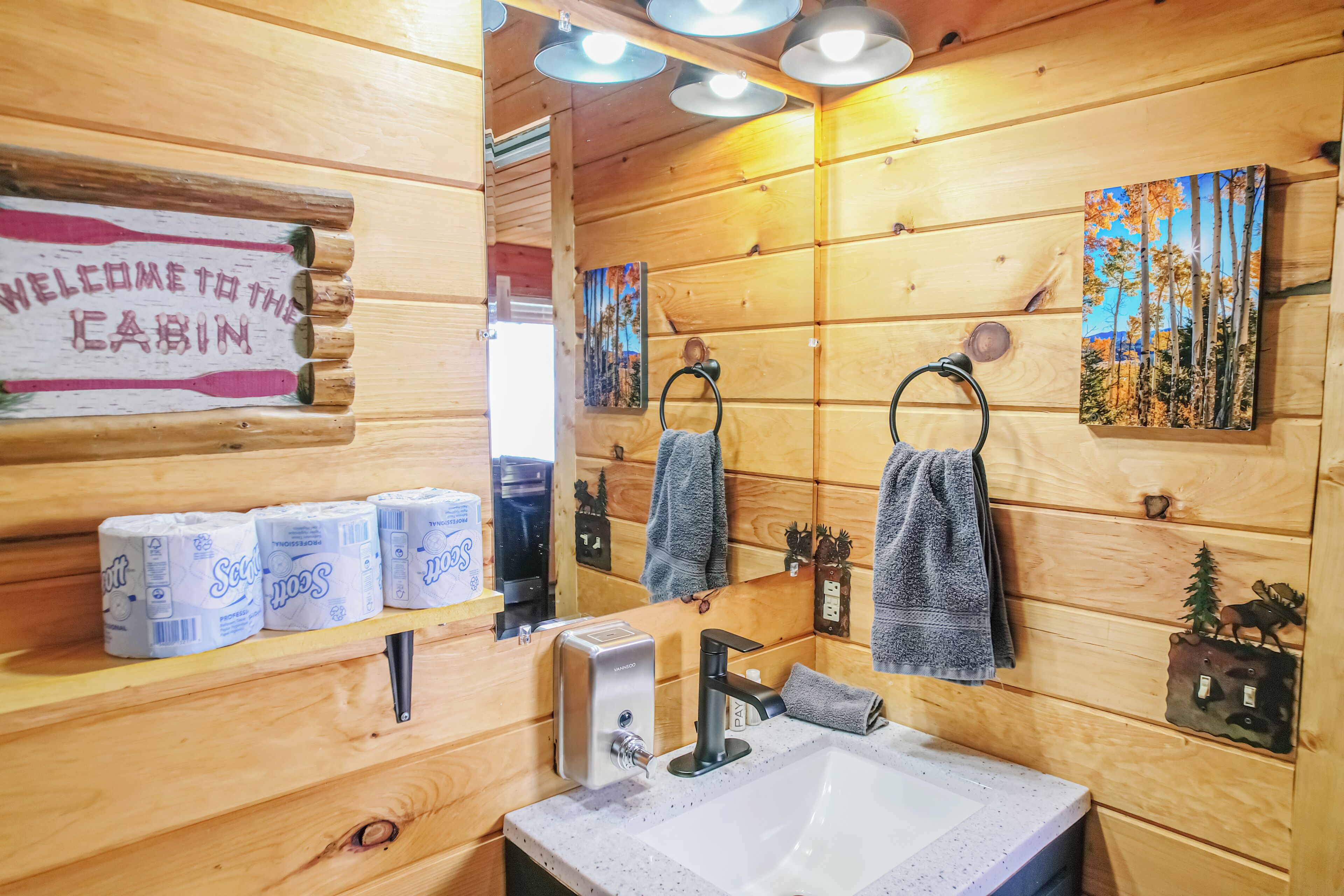 Bagno di una baita in legno, con lavandino, carta igienica, asciugamani e un cartello di benvenuto.