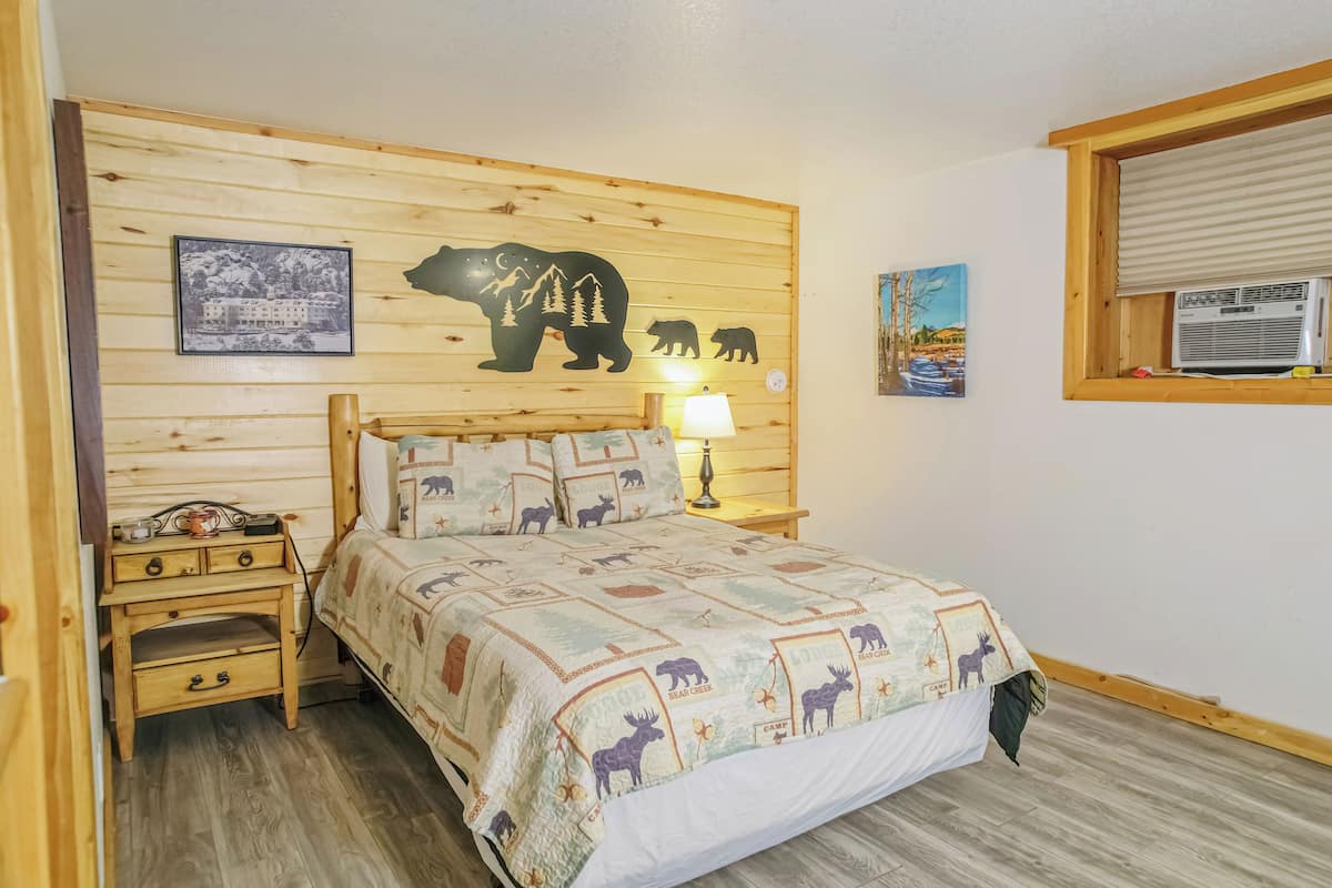 A bedroom with a wooden headboard, a bed with a bear-themed quilt, a nightstand with a lamp, and a window with an air conditioner.