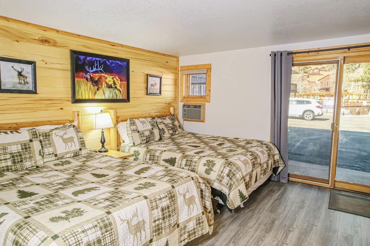 A room with two beds, a wooden wall, a painting, and a window with a view of a car outside.