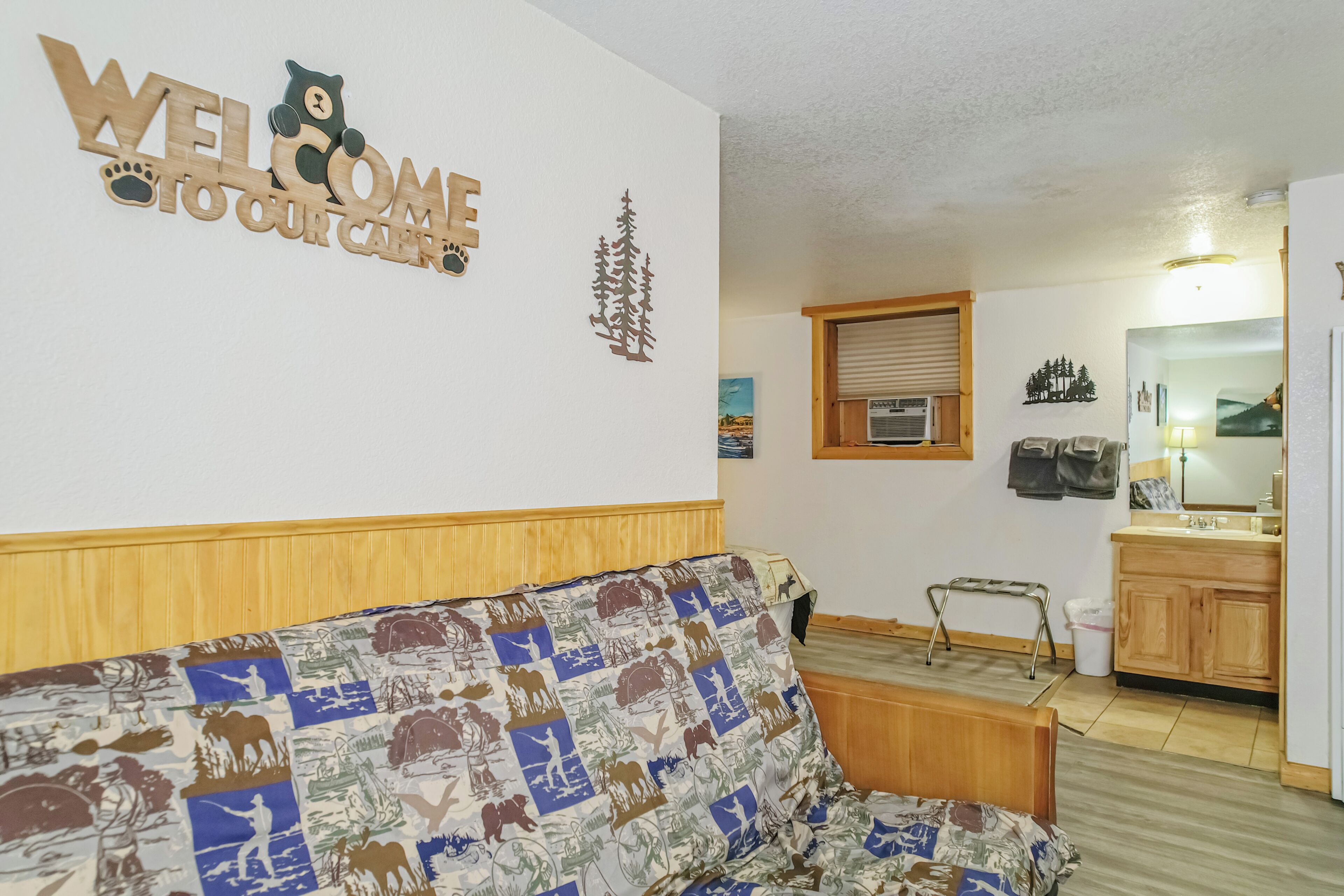 Ein Wohnzimmer mit einer Couch, einem hölzernen Bettgestell, einer Wand mit der Aufschrift 'WELCOME TO OUR CAMP', einer kleinen Küchenzeile und einem Fenster mit Jalousien.