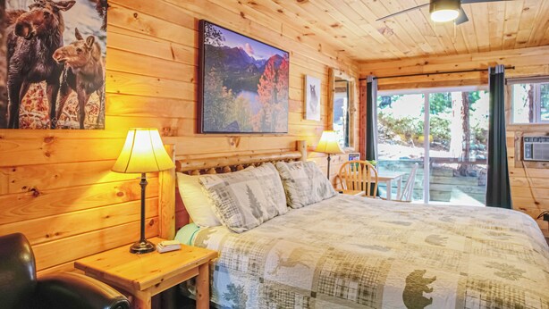 A wooden cabin room with a bed, a lamp, a chair, and a painting of moose. - Nicky's Resort (Estes Park)