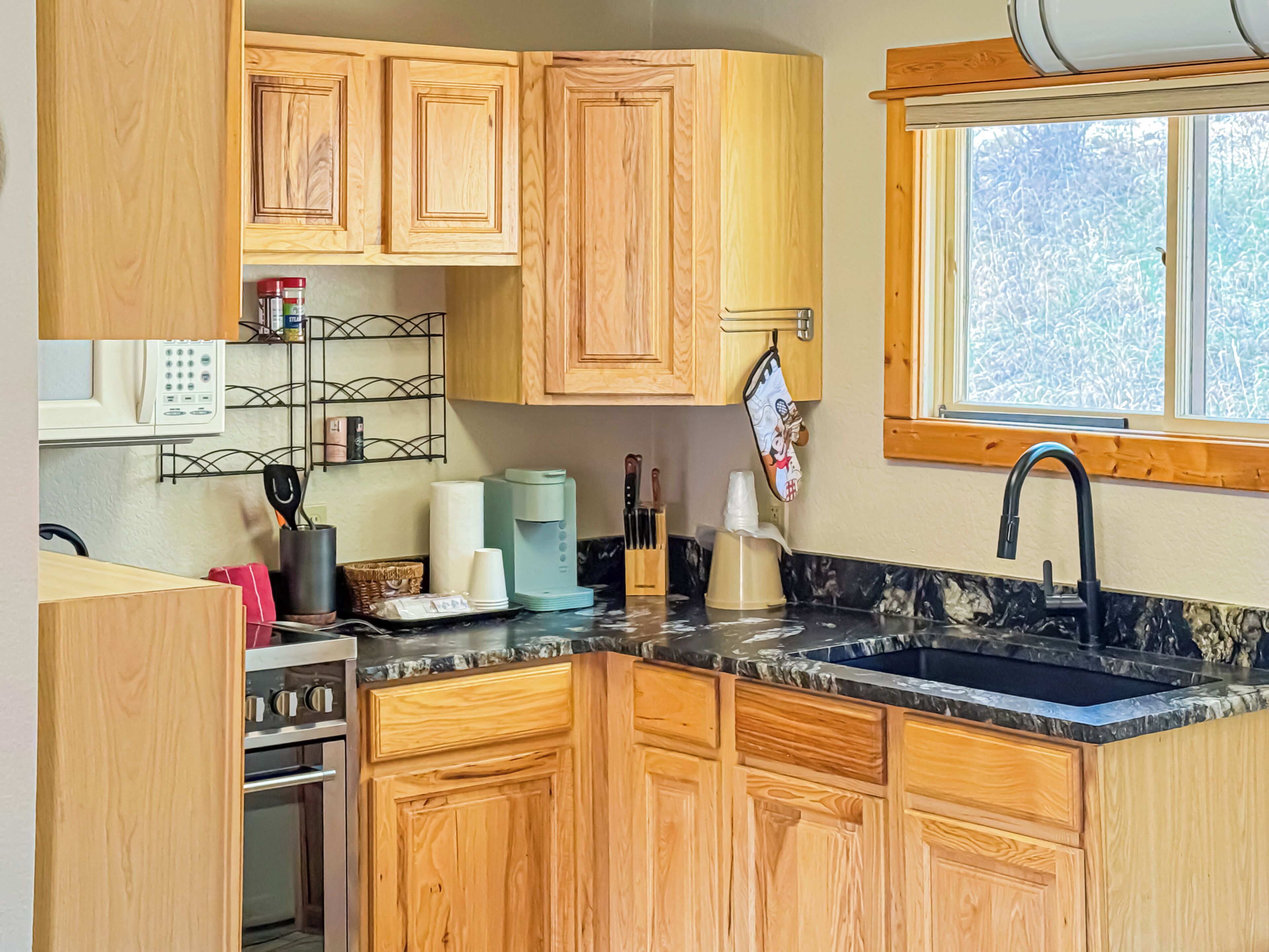 A compact kitchen with wooden cabinets, a built-in oven, a coffee maker, and a window with a view of trees.