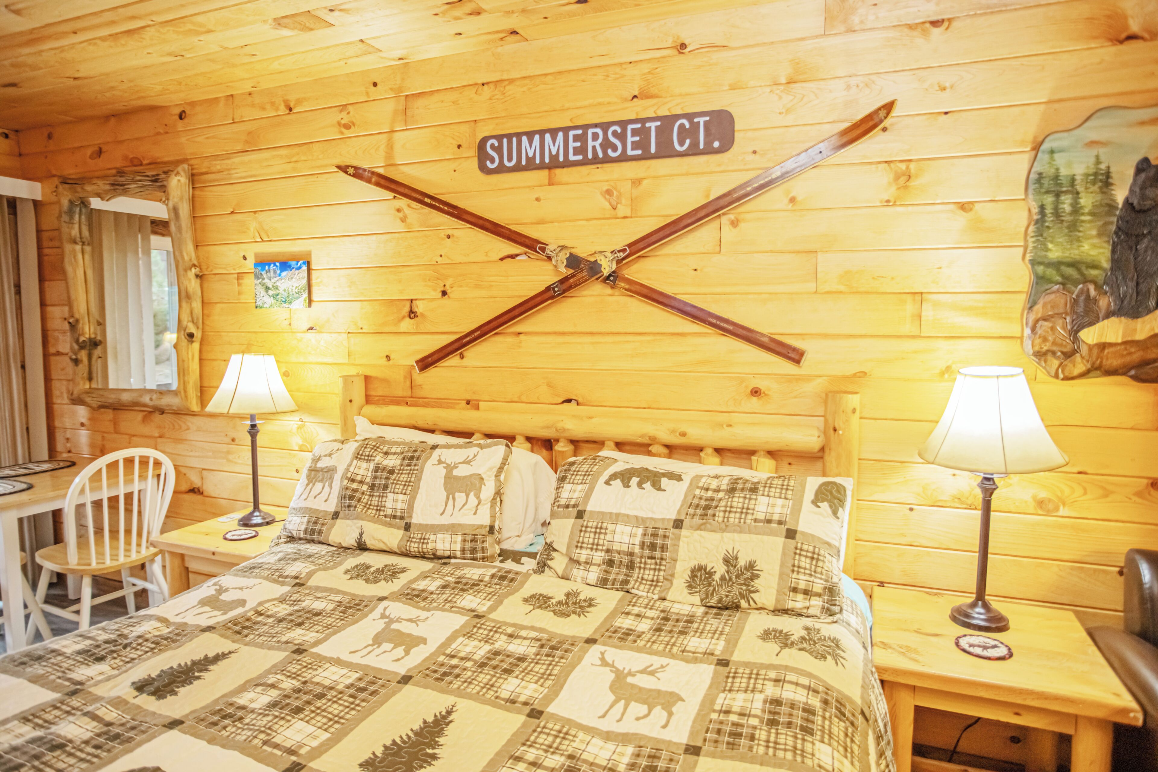 A wooden cabin room with a bed, two lamps, a chair, and a wall sign reading 'SUMMERSET CT.'