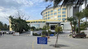 A hotel entrance with a signboard, palm trees, and a parking area.