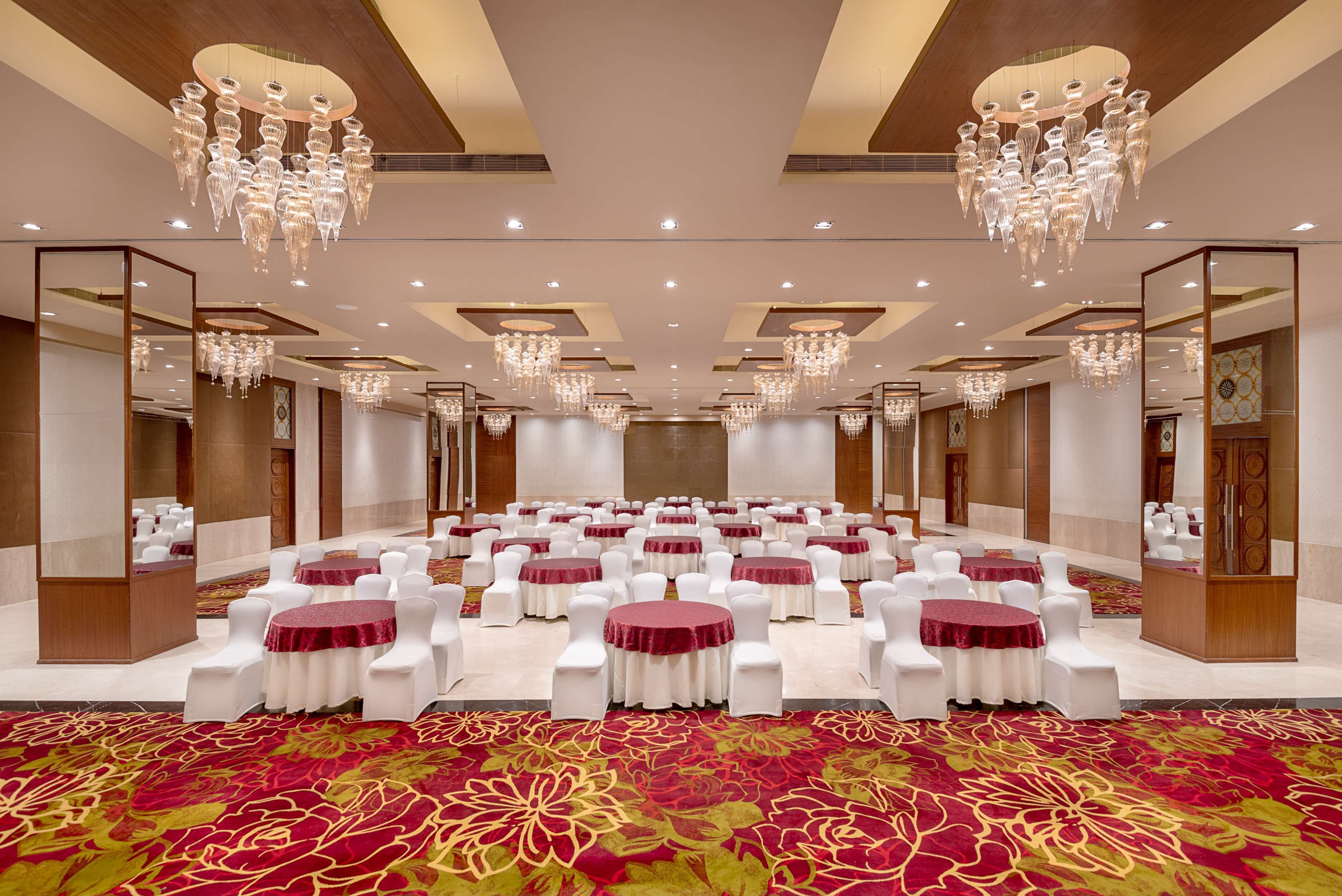 A large conference room with round tables, white chairs, and red tablecloths, featuring chandeliers and a patterned carpet.