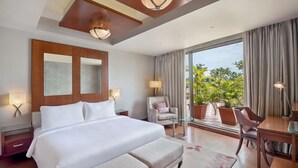 A hotel room with a large bed, a desk, a chair, a lamp, and a view of greenery outside.