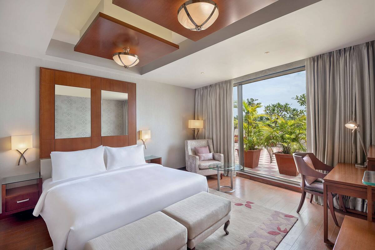A hotel room with a large bed, a desk, a chair, a lamp, and a view of greenery outside.