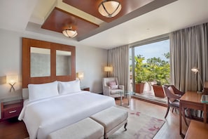 A hotel room with a large bed, a desk, a chair, a lamp, and a view of greenery outside.