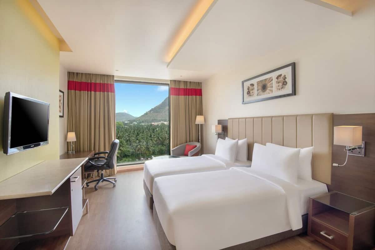 A hotel room with two beds, a desk, a TV, and a view of mountains.