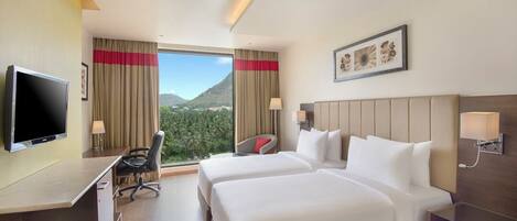 A hotel room with two beds, a desk, a TV, and a view of mountains.