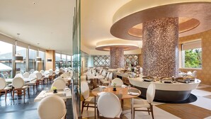 A modern restaurant with round tables and white chairs, featuring a central mosaic column and a view of greenery outside.