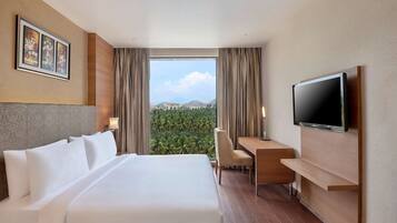 Ein Hotelzimmer mit einem großen Bett, einem Schreibtisch, einem Sessel, einem Fernseher und Blick ins Grüne durch das Fenster.