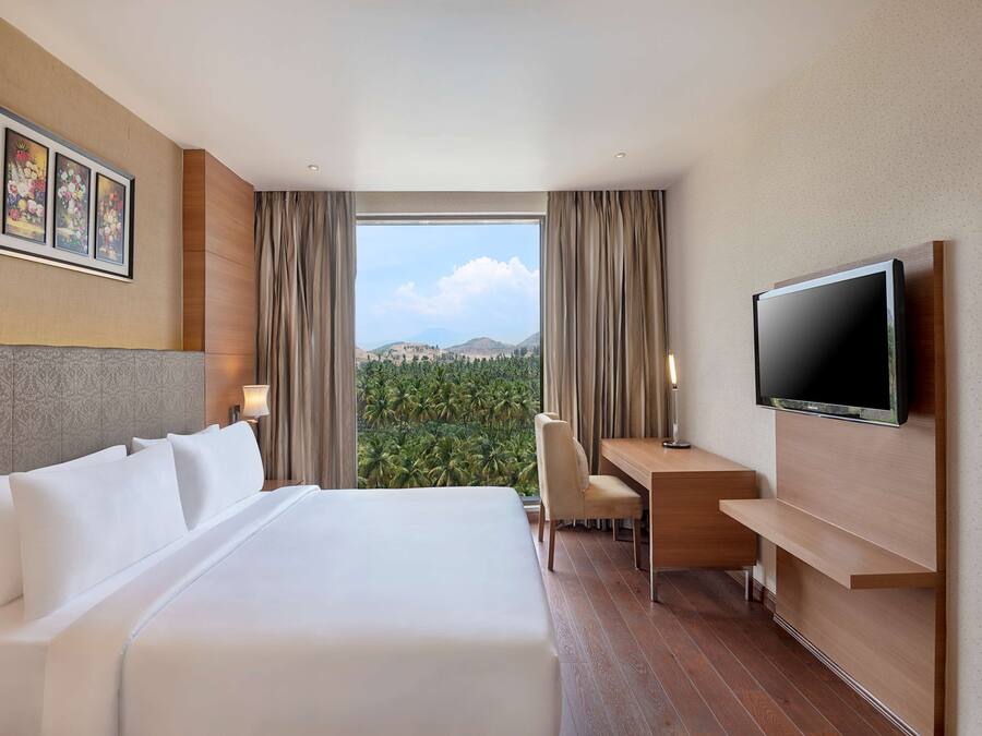 Ein Hotelzimmer mit einem großen Bett, einem Schreibtisch, einem Sessel, einem Fernseher und Blick ins Grüne durch das Fenster.