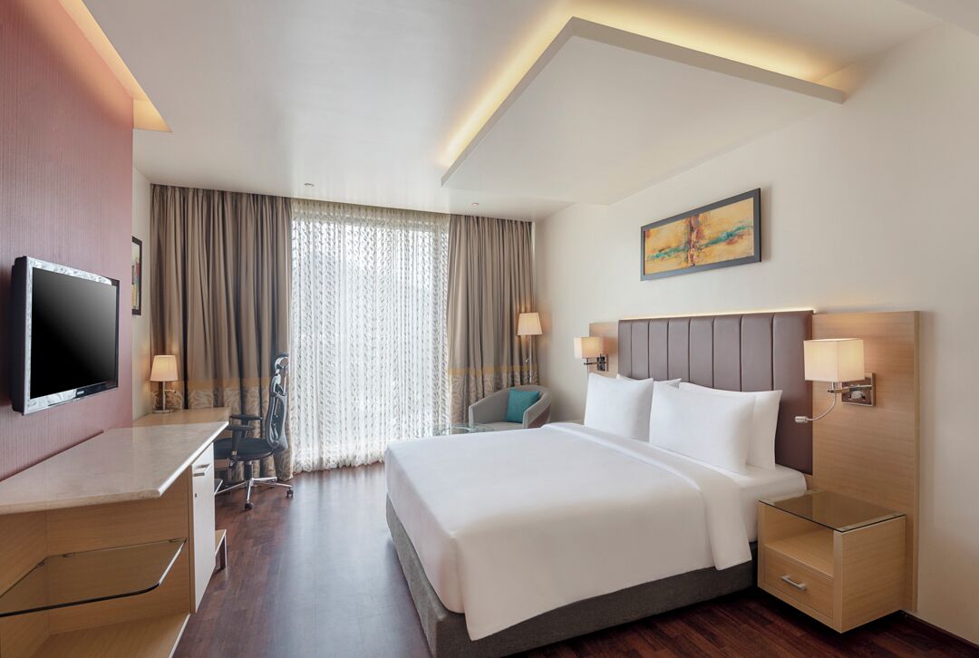 A modern hotel room with a large bed, bedside tables, a desk, a chair, and a TV.