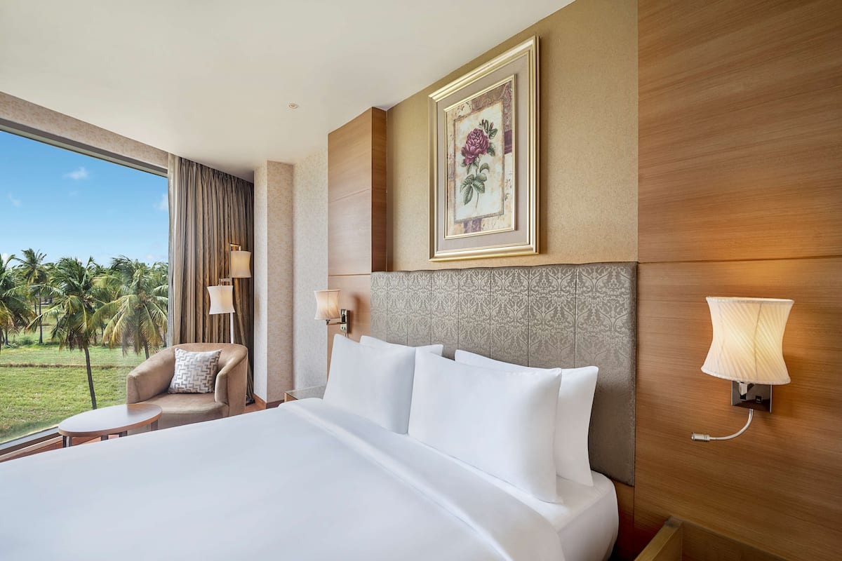A hotel room with a large bed, a sitting area with a chair and table, a view of palm trees, and a framed artwork on the wall.