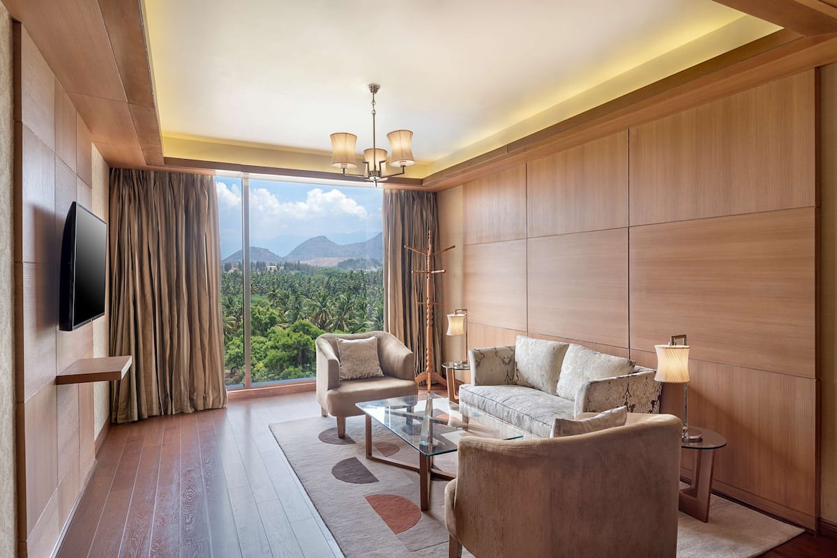 A modern hotel room with a large window, a flat-screen TV, a sofa, two armchairs, a coffee table, and a lamp.