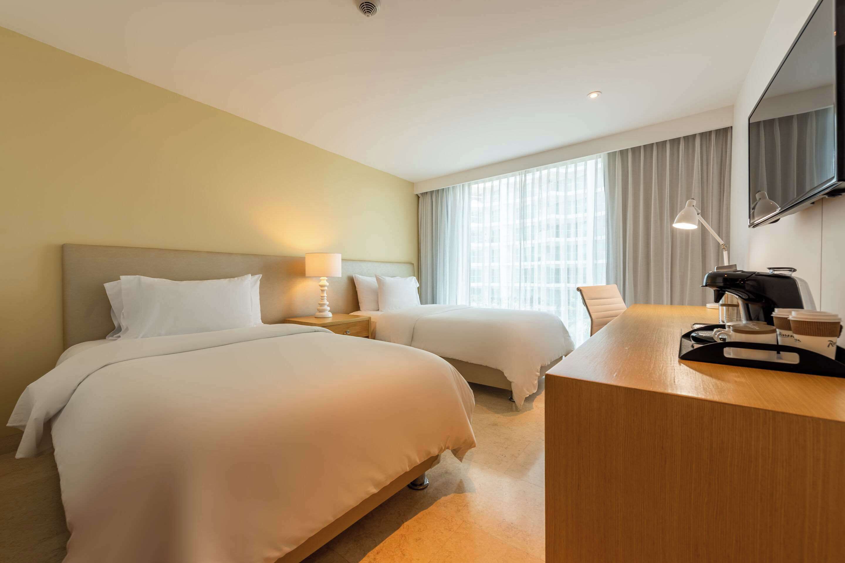 A hotel room with two beds, a desk, a TV, and a window with curtains.