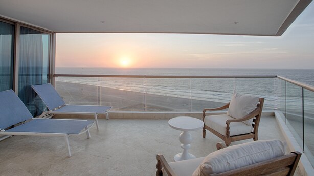 Un balcón con vistas a la puesta de sol sobre el océano, amueblado con sillones y una mesa.