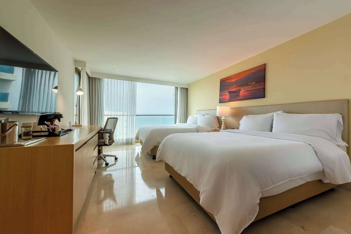 Une chambre d’hôtel avec deux lits, un bureau, une chaise et une grande fenêtre donnant sur la mer.